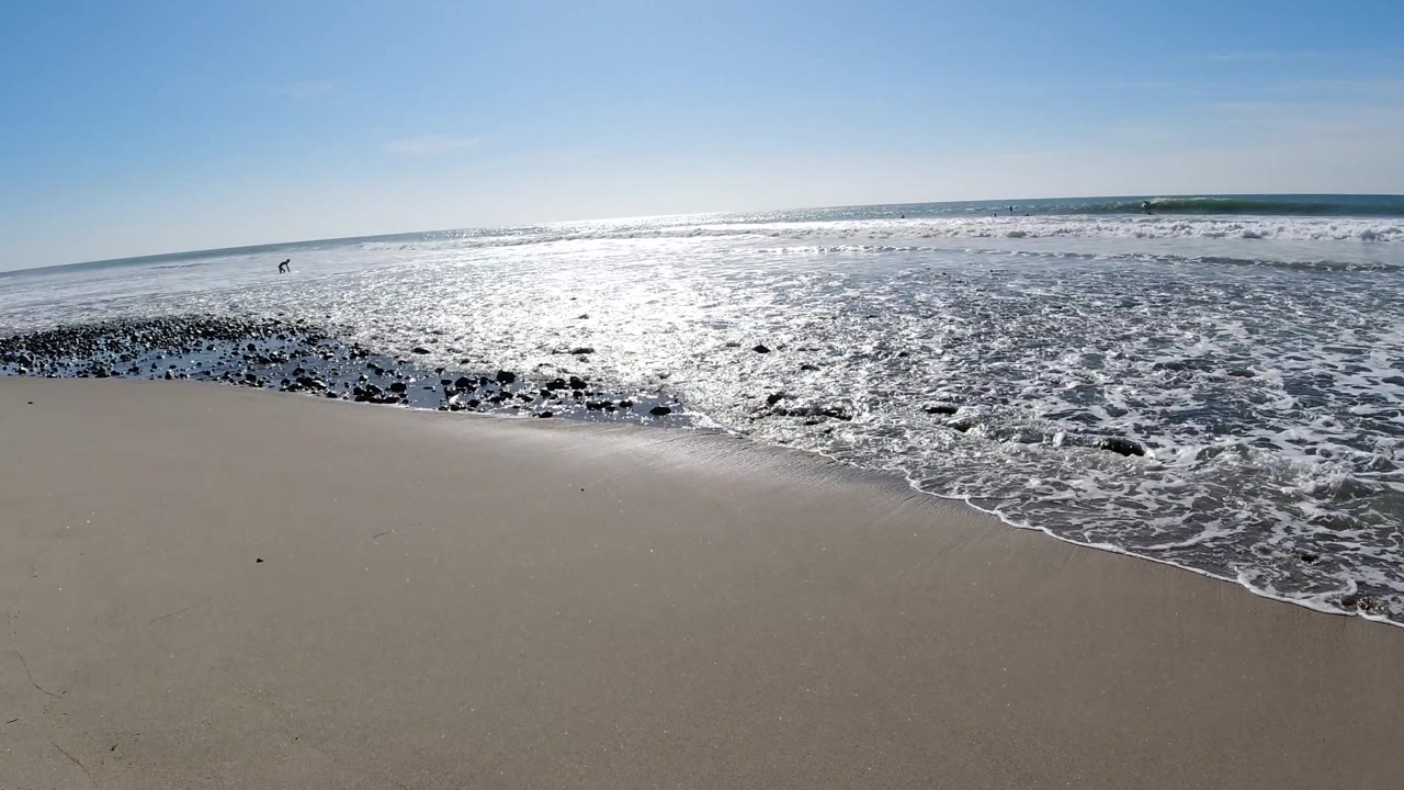 Sandy beach at Trestles Beach, California, with a diagonal line of white surf and dark rocks in the midground, sun-reflected ocean, and no visible people