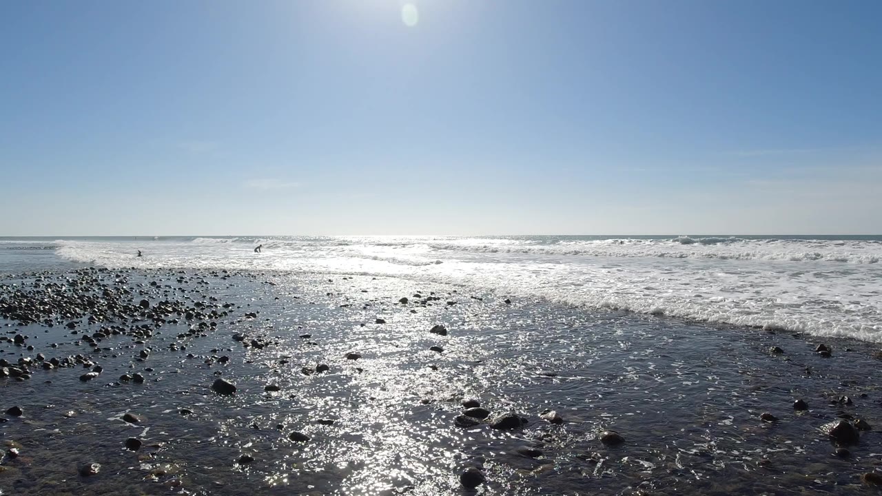 Sun-lit rocky shoreline at Trestles Beach, California, with dark wet pebbles in the foreground, shallow wave wash spreading across the sand, ocean horizon and clear blue sky beyond; no visible people