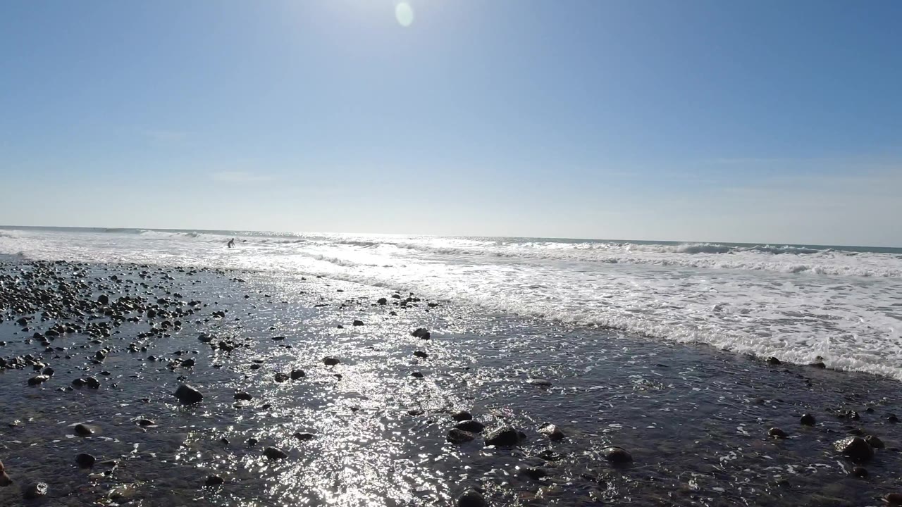 Trestles Beach, California rocky shoreline at 0:02, scattered dark pebbles and rocks on glistening wet sand in foreground, ocean wave water in mid-ground, and blue sky above; no visible people