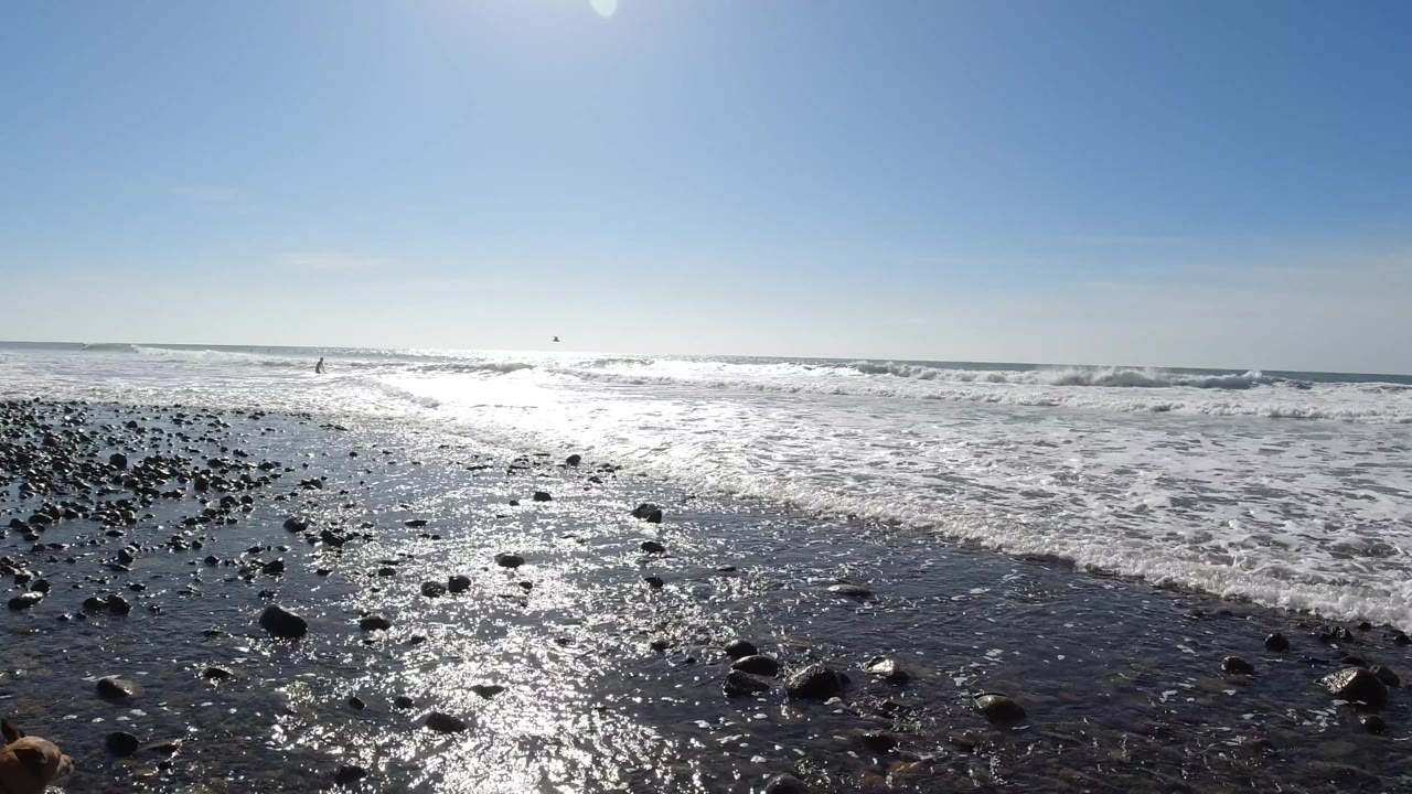 Trestles Beach, California shoreline at the midpoint, with dark wet pebbles and rocks in the foreground, shallow wave water spreading across the beach, and the ocean horizon under a clear blue sky; no visible people