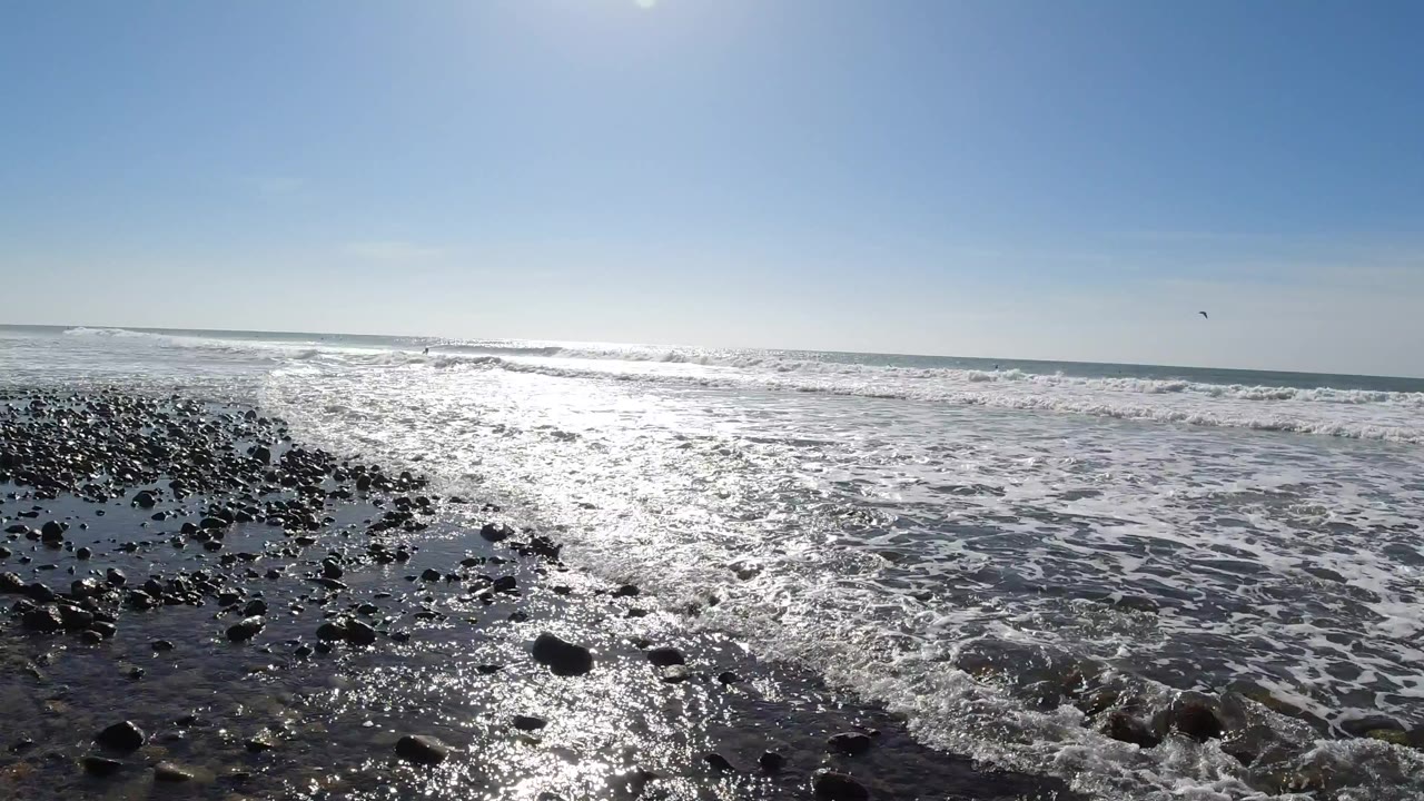 Low ground-level view of dark wet rocks and rounded pebbles at Trestles Beach, California, with shallow foamy seawater washing over them, the sun-lit ocean horizon behind, and a cloudless blue sky; no people are visible.