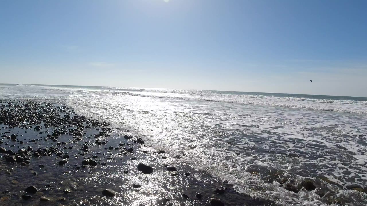 Ground-level view of dark rocks and wet pebbles at Trestles Beach, California, with shallow foamy seawater shifting around the stones and the sun reflecting off the wet shore; no people are visible.
