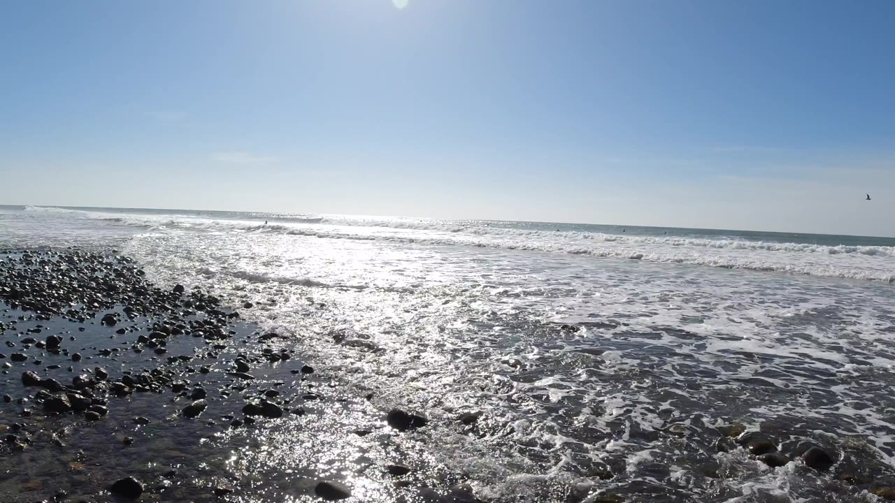 Low-angle view of wet dark rocks and pebbles at Trestles Beach, California, with shallow foamy seawater and the ocean horizon lit by direct overhead sun; no people are visible.