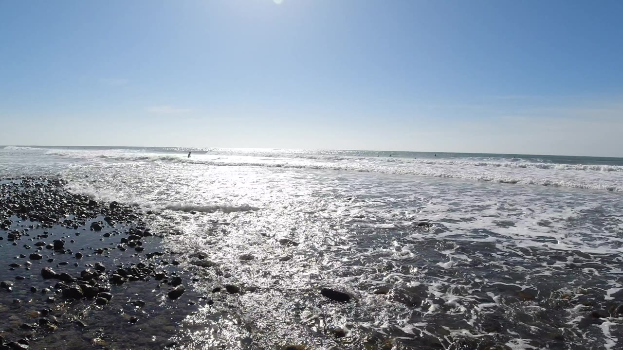 Wet dark rocks in the foreground with shallow foamy water, the sunlit ocean extending to the horizon, and clear blue sky; a tiny indistinct shape is visible at the far horizon.