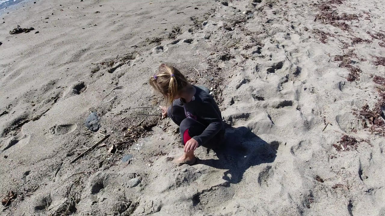 The child remains kneeling in thick sand at Trestles Beach, California, with footprints and dry seaweed surrounding the play area.