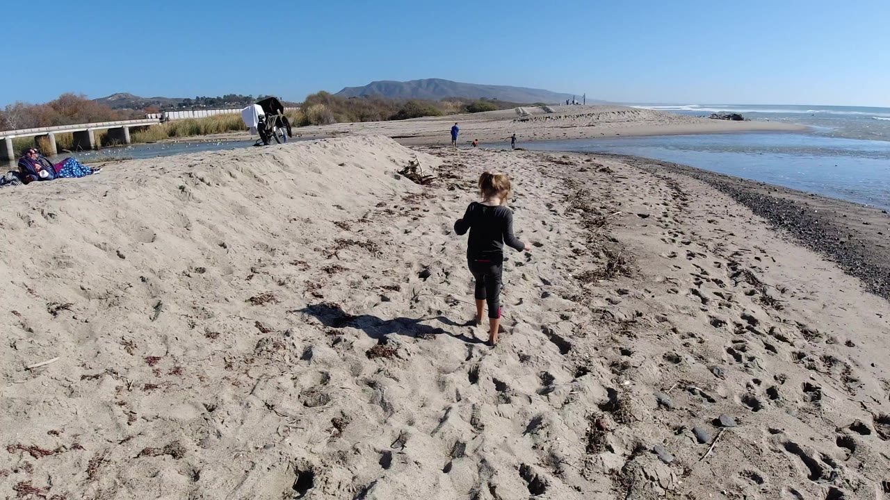 At Trestles Beach, California, a child in dark clothing walks away from the camera across pale sand toward the shallow ocean water, with beach gear and a few people in the distance.