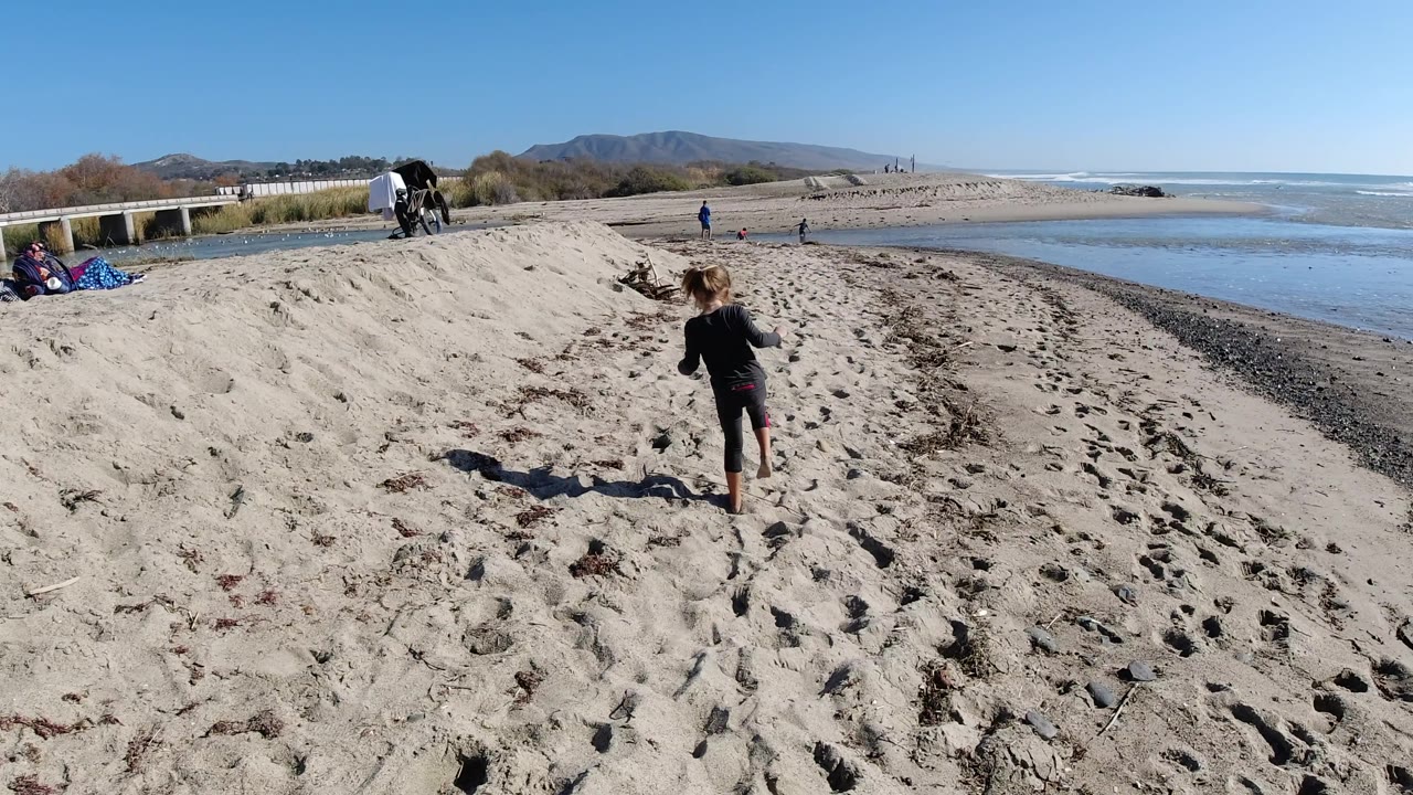 At Trestles Beach, California, the child is slightly farther down the sandy slope, with a dark shadow on the sand, shallow blue water to the right, and sparse beachgoers near the shoreline.