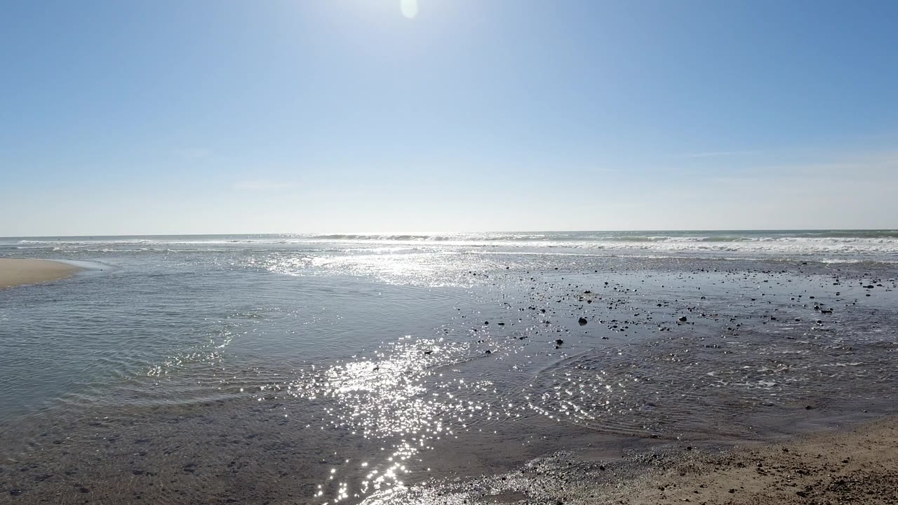 Low-angle wide shot of the sunlit tidal flat at Trestles Beach, California, with reflective shallow water, scattered dark pebbles on wet sand, and clear blue sky; no visible people