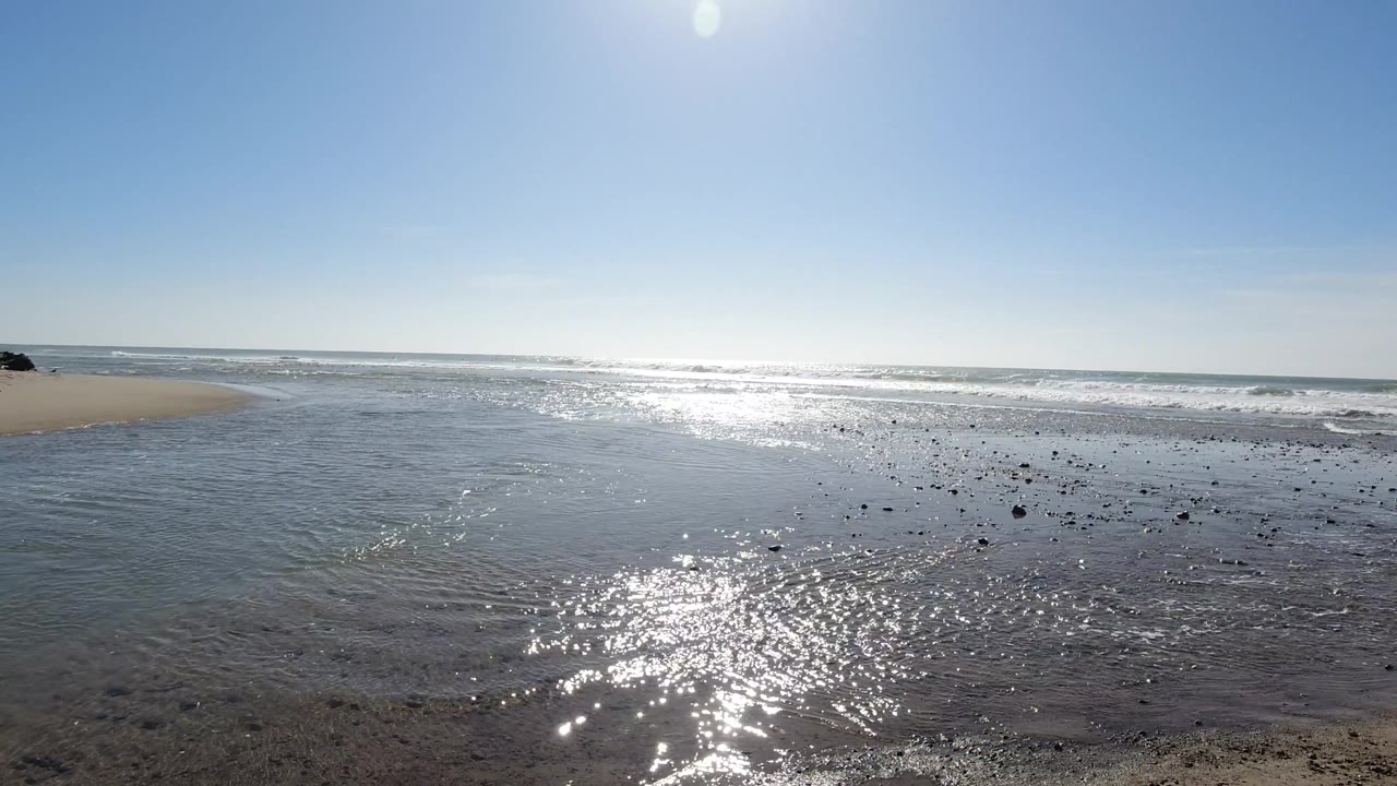 Wide low-angle view of flat wet sand at Trestles Beach, California, with sun reflection on tidal water and calm ocean horizon under a clear blue sky; no visible people