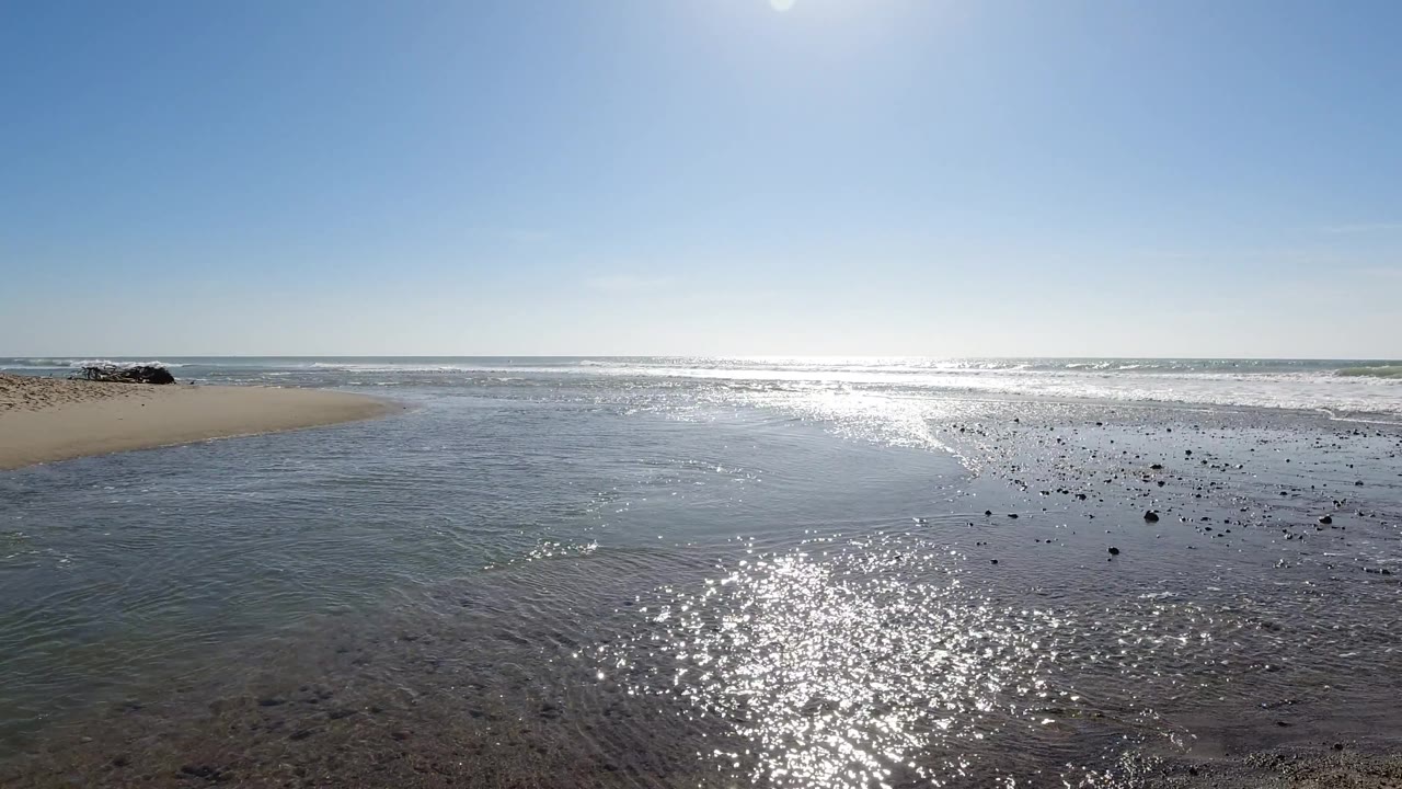 At two seconds, the Trestles Beach composition holds wide and low-angle, with the specular highlight field persisting and dark objects appearing in a slightly denser cluster toward the left midground compared to the opening frame; no people visible.