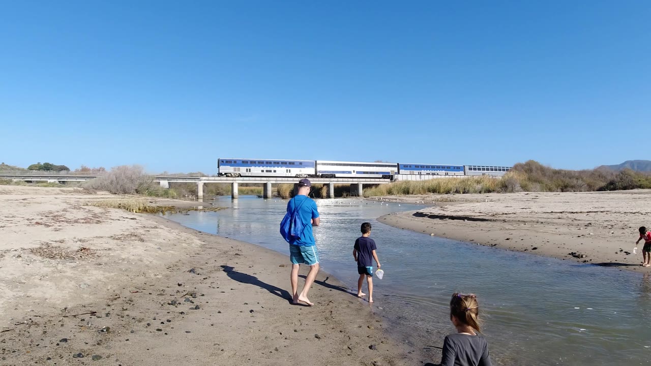 At Trestles Beach, California, the adult in blue and two children remain near the river edge as another child stands on the opposite sandbar and the train continues across the bridge.