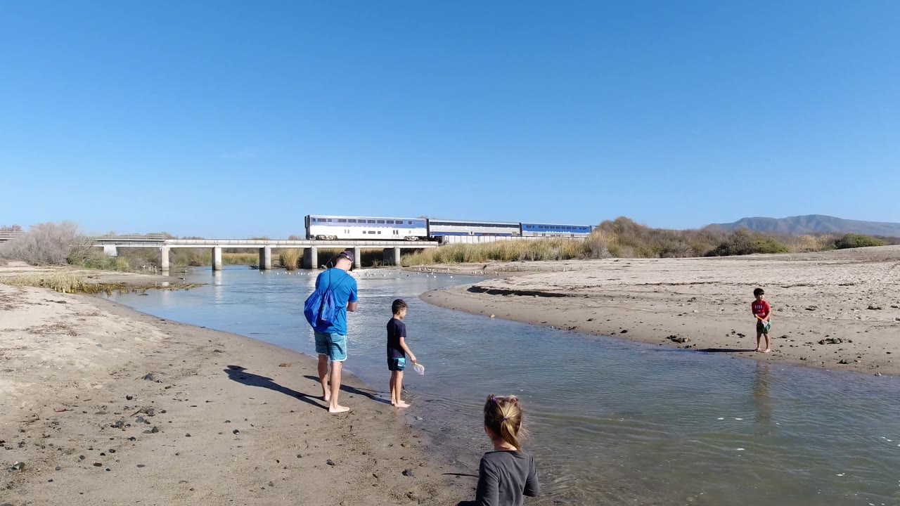 At Trestles Beach, California, children are spaced across shallow water and sand, with the adult near the left bank and the train still visible on the bridge in the background.