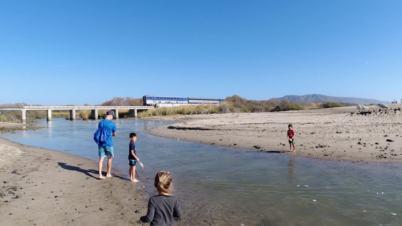 Family group by shallow water with train and low bridge in the distance.