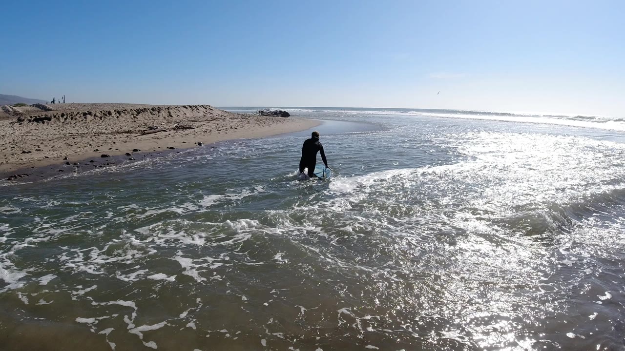 One second in at Trestles Beach, the surfer stands slightly more upright at the shoreline, with foamy surf washing around them and sun-bright wet sand filling most of the lower frame; the rock jetty is faintly visible in the background, and a person is visible.