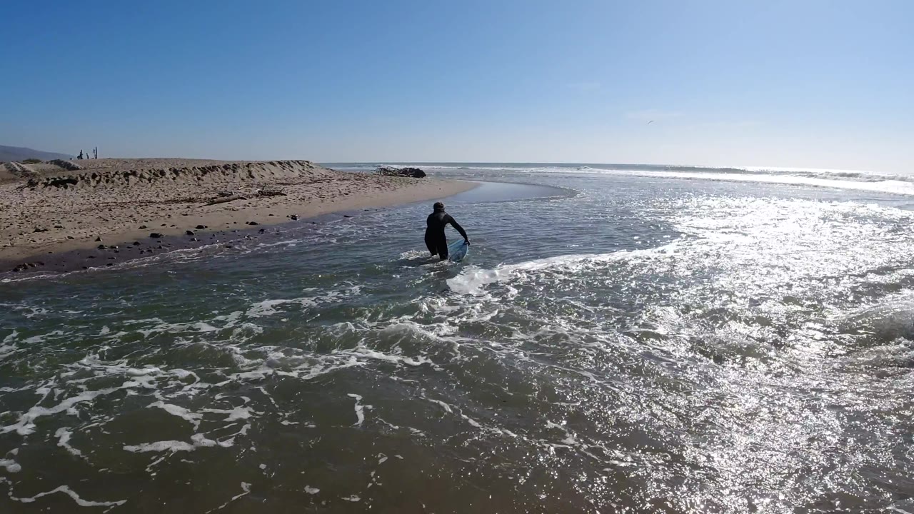 At two seconds at Trestles Beach, the surfer in a black wetsuit stands at the edge of the shallow surf, with the rock breakwater now more clearly defined in the right background and strong sun glare still dominating the wet sand in the foreground; a person is visible.
