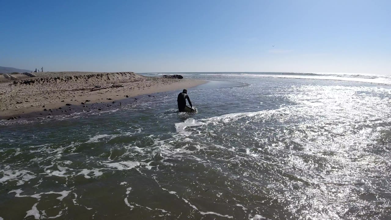 Aerial shot of a surfer in a black wetsuit walking toward the dry beach, with shallow foamy water in the foreground.
