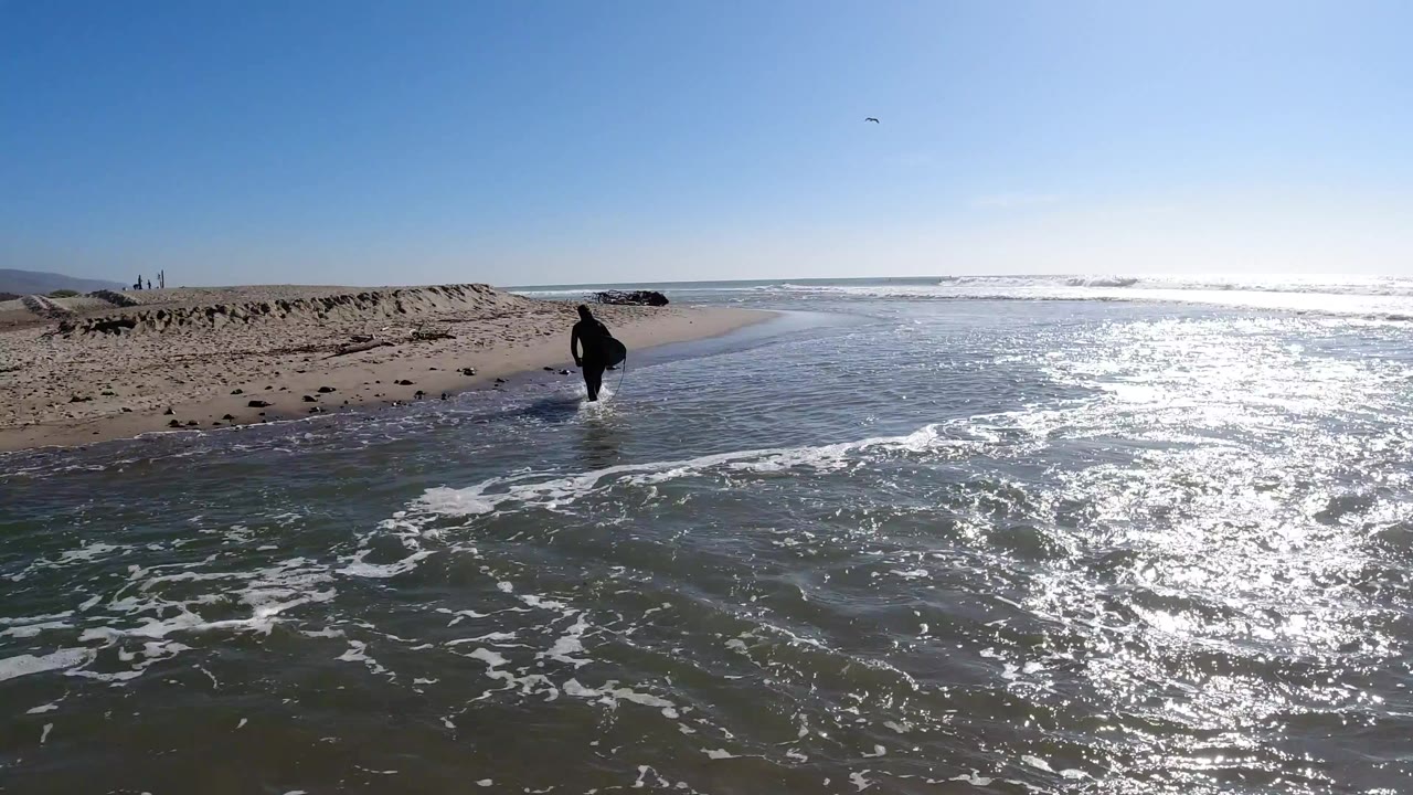 At Trestles Beach, California, a man in dark clothing walks through shallow water near the sand as bright sun reflects across the ocean surface.