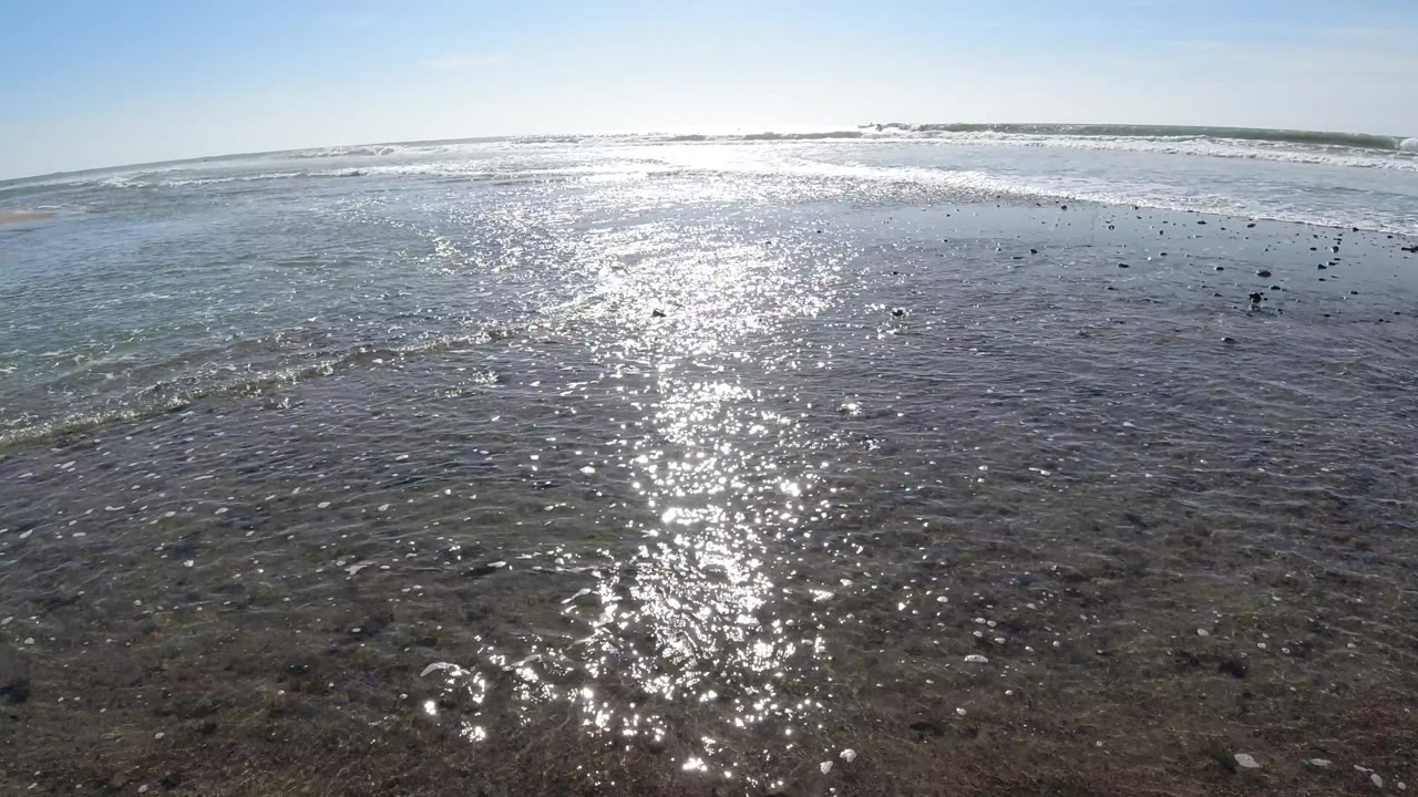 At Trestles Beach, California, sunlight scatters across a broad shallow water surface with darker submerged patches and a band of breaking waves in the background, with no visible people.