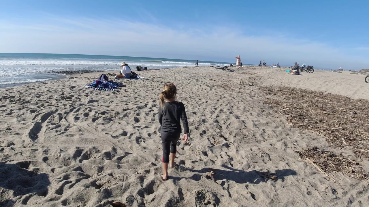 At Trestles Beach, California, a young girl in dark clothes walks away from camera across rippled sand toward the surf, with towels, seated beachgoers, and the ocean horizon ahead.
