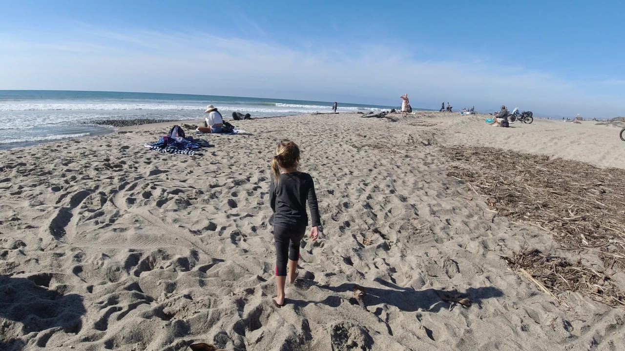 Girl continues across sandy beach toward low waves and scattered people.