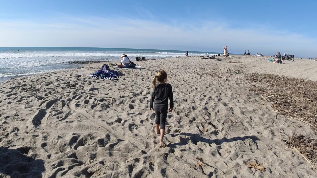 At Trestles Beach, California, the child is slightly farther forward on the sand, with a long shadow beside her, surf rolling in from the left, and small groups of beachgoers spread across the background.