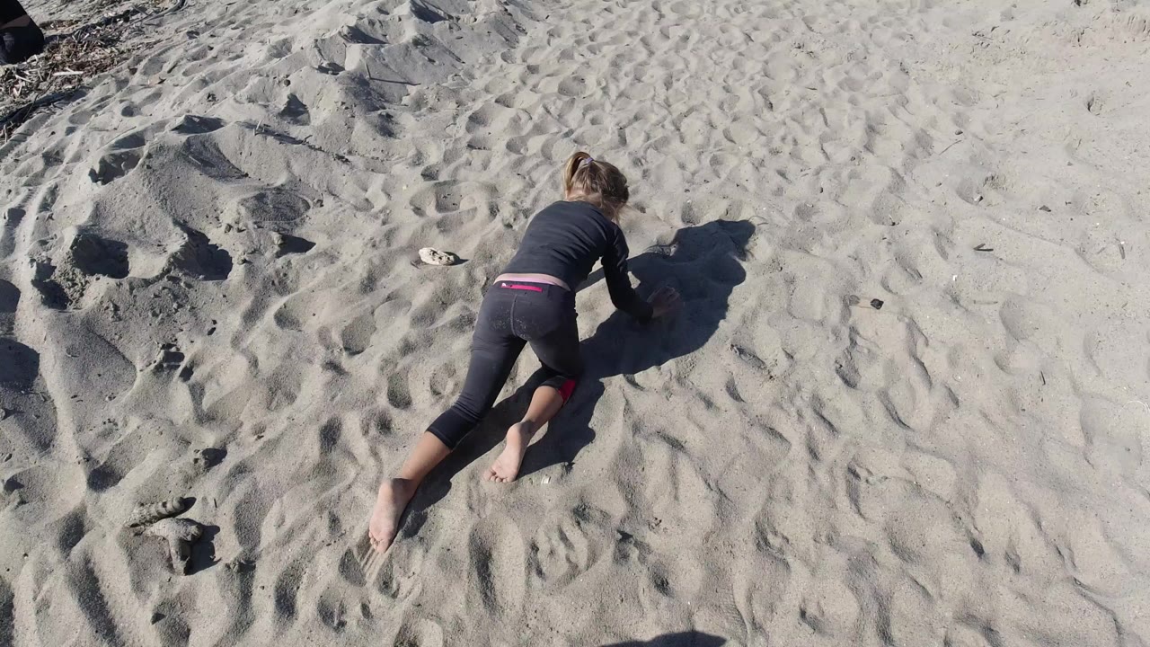 Overhead frame at Trestles Beach, California showing a child in dark clothing crawling across pale sand beside low sand mounds and footprints.
