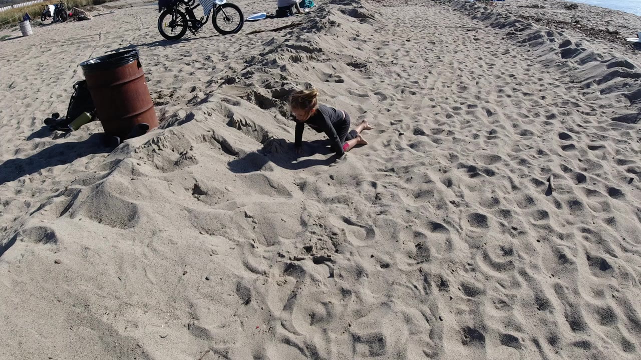At Trestles Beach, California, a young girl plays low on the sand beside a rust-colored barrel, with bicycles and scattered beach items near the top of frame.