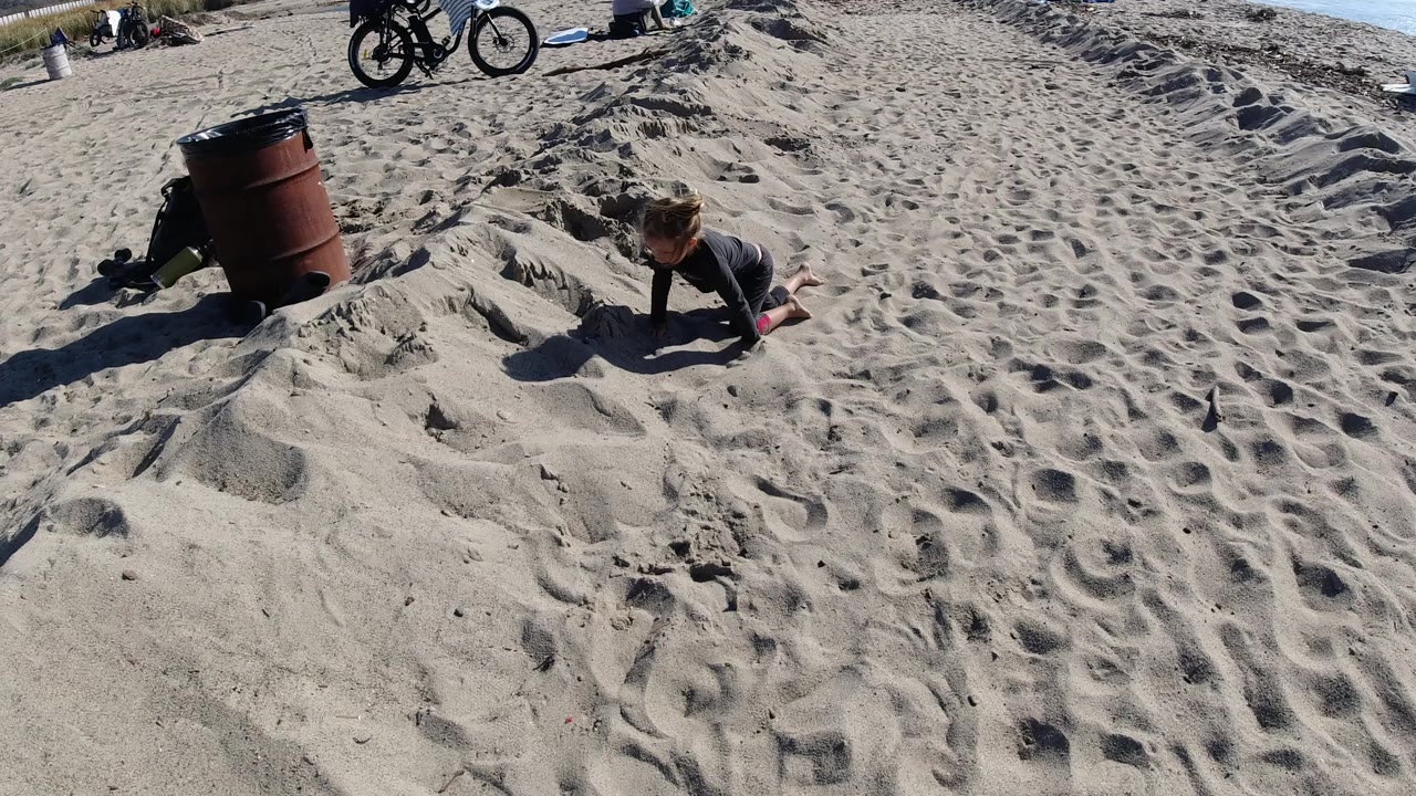 At Trestles Beach, California, the girl is crouched closer to the sand, with hard sunlight throwing a long shadow and beach bicycles still visible in the background.