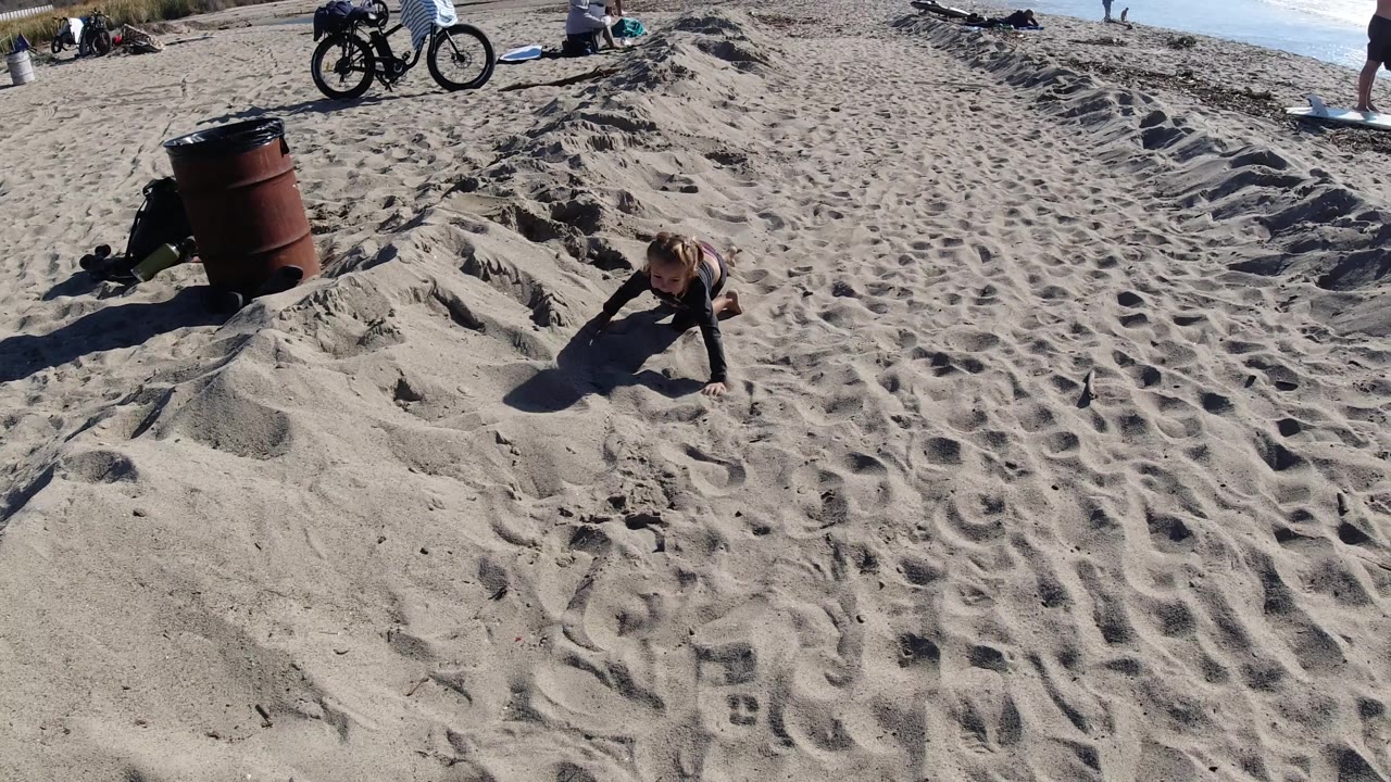 At Trestles Beach, California, the girl crawls across rippled sand while bicycles, towels, and small figures near the waterline sit along the upper frame.