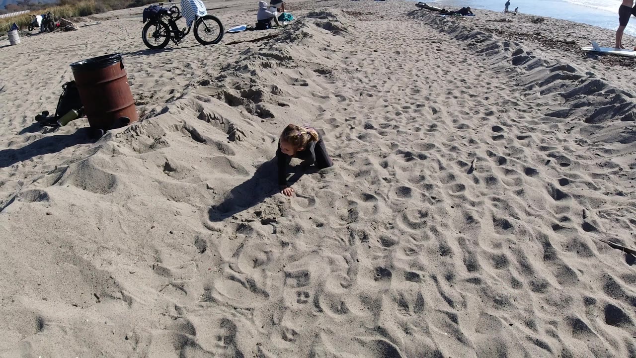Girl leaning forward on sand with barrel and bicycles behind her.