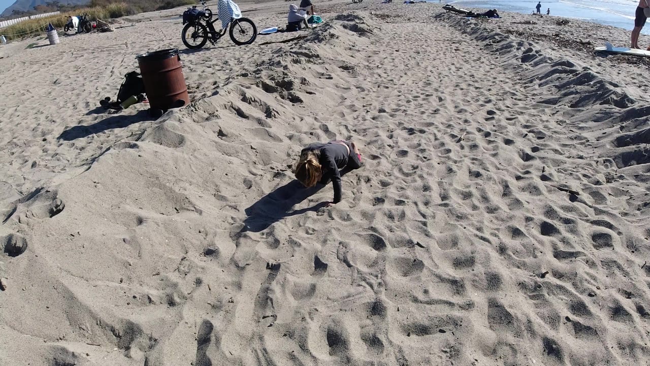 Adult female crawls on all fours across patterned beach sand near a trash barrel.