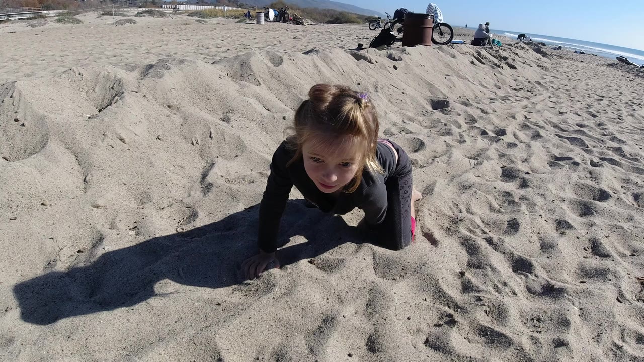A young child crawls toward the camera on sandy Trestles Beach, California, with long shadows, distant beachgoers, bicycles, blue sky, and ocean in the background.