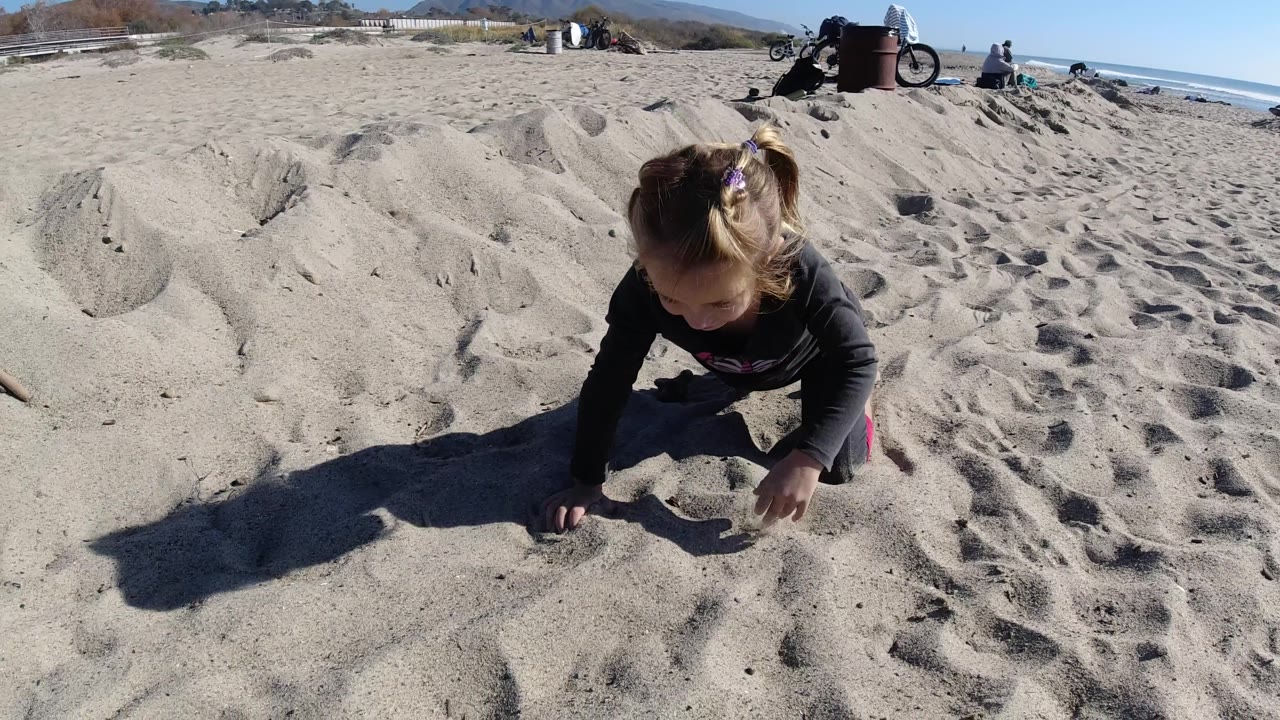 The child looks down and places both hands in the sand at Trestles Beach, California, with footprints, small sand ridges, distant people, and ocean behind.