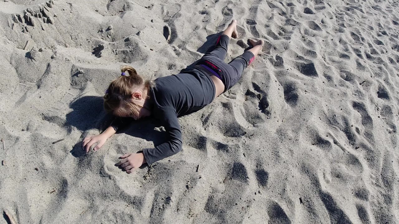 Overhead view of a young girl lying on pale rippled sand at Trestles Beach, California, with her arms forward and bare feet visible in bright sunlight.