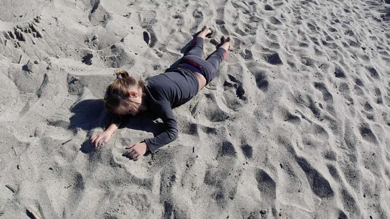 Overhead frame of the young girl on her stomach at Trestles Beach, California, with both hands near the sand and a field of footprints filling the right side.