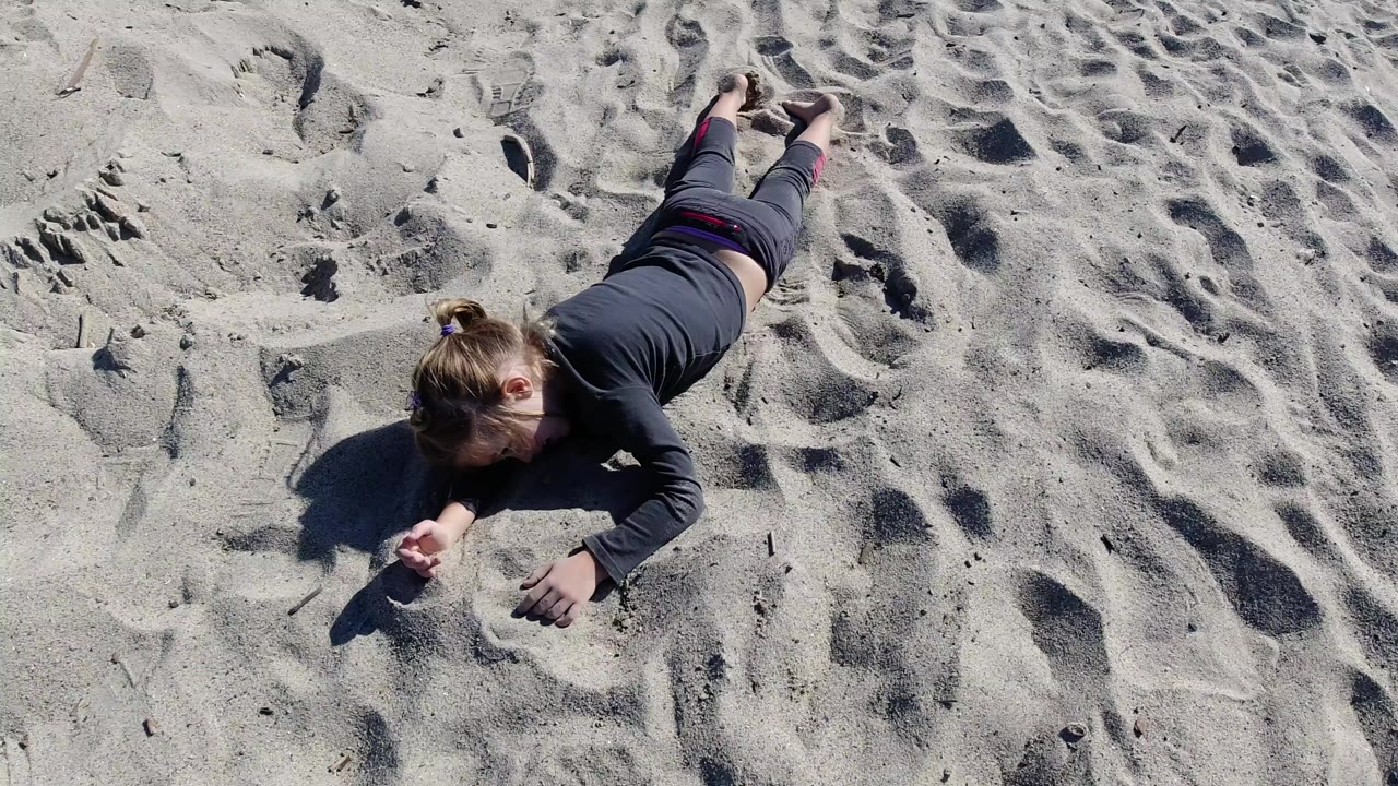 Overhead view at Trestles Beach, California, of an adult lying face down on pale rippled sand with one arm bent near the foreground and strong sunlight casting a left-side shadow.