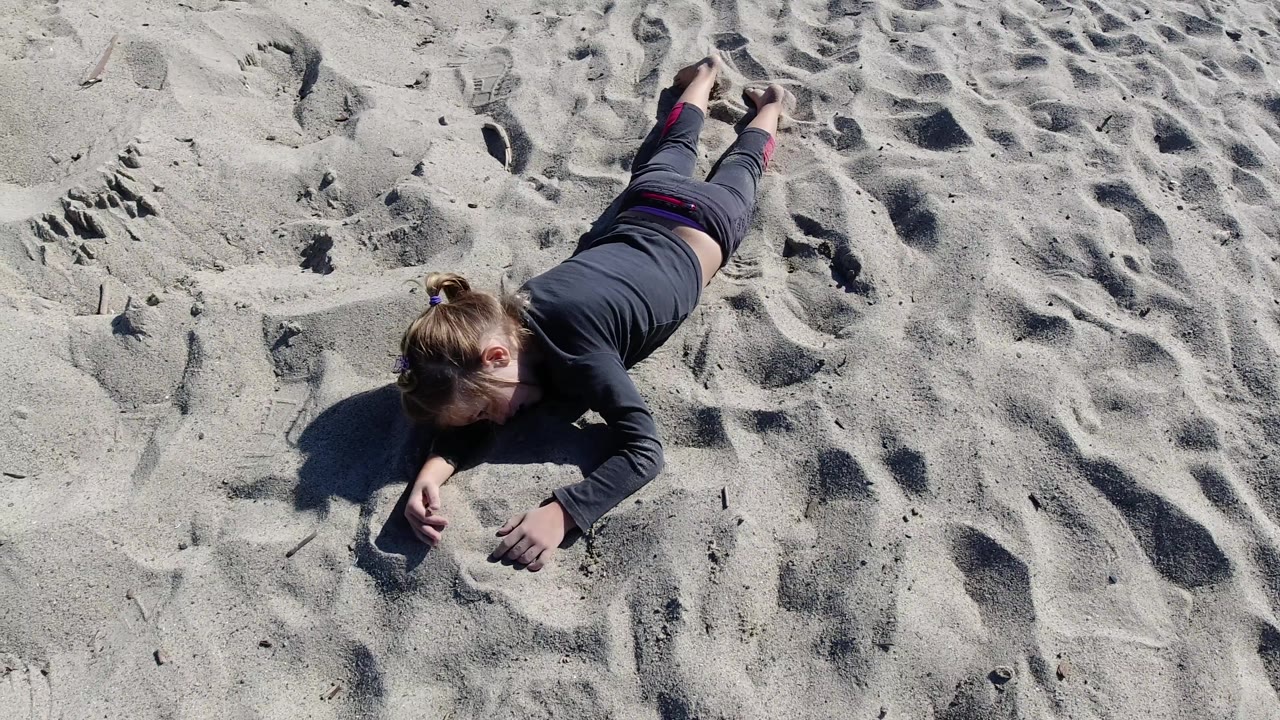 At Trestles Beach, California, the adult remains stretched out on the sand as one hand lifts slightly, with bright sun emphasizing ridges and depressions in the surrounding beach surface.