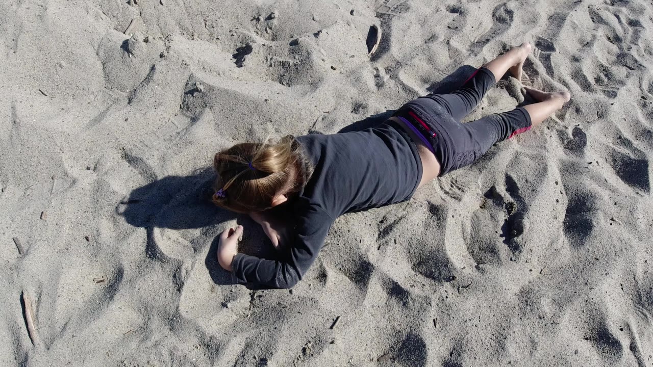 Overhead view at Trestles Beach, California, showing a young girl lying diagonally on pale sand with footprints and a long shadow to her left.