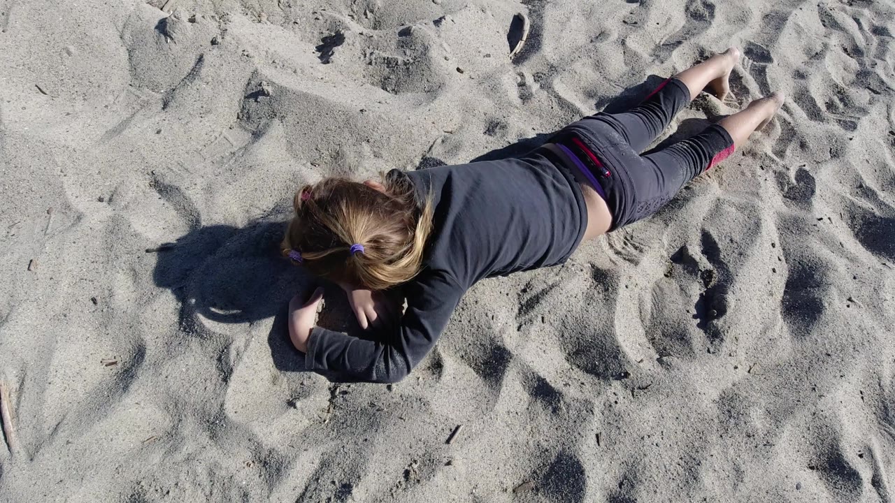 The child remains stretched across the frame at Trestles Beach, California, with her ponytail visible, bare feet at the upper right, and sunlit sand filling the background.