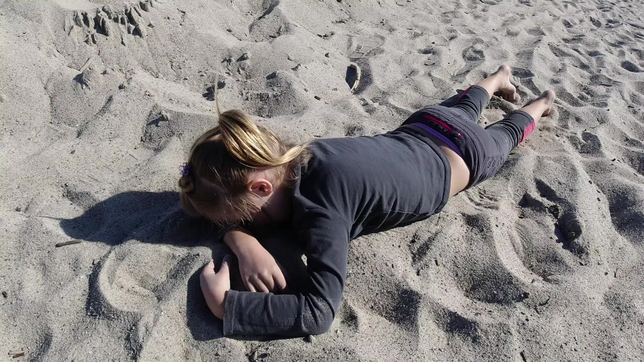 At Trestles Beach, California, a young girl in dark clothing lies face down on textured sand with her arms folded near her head and bare feet extended behind her.