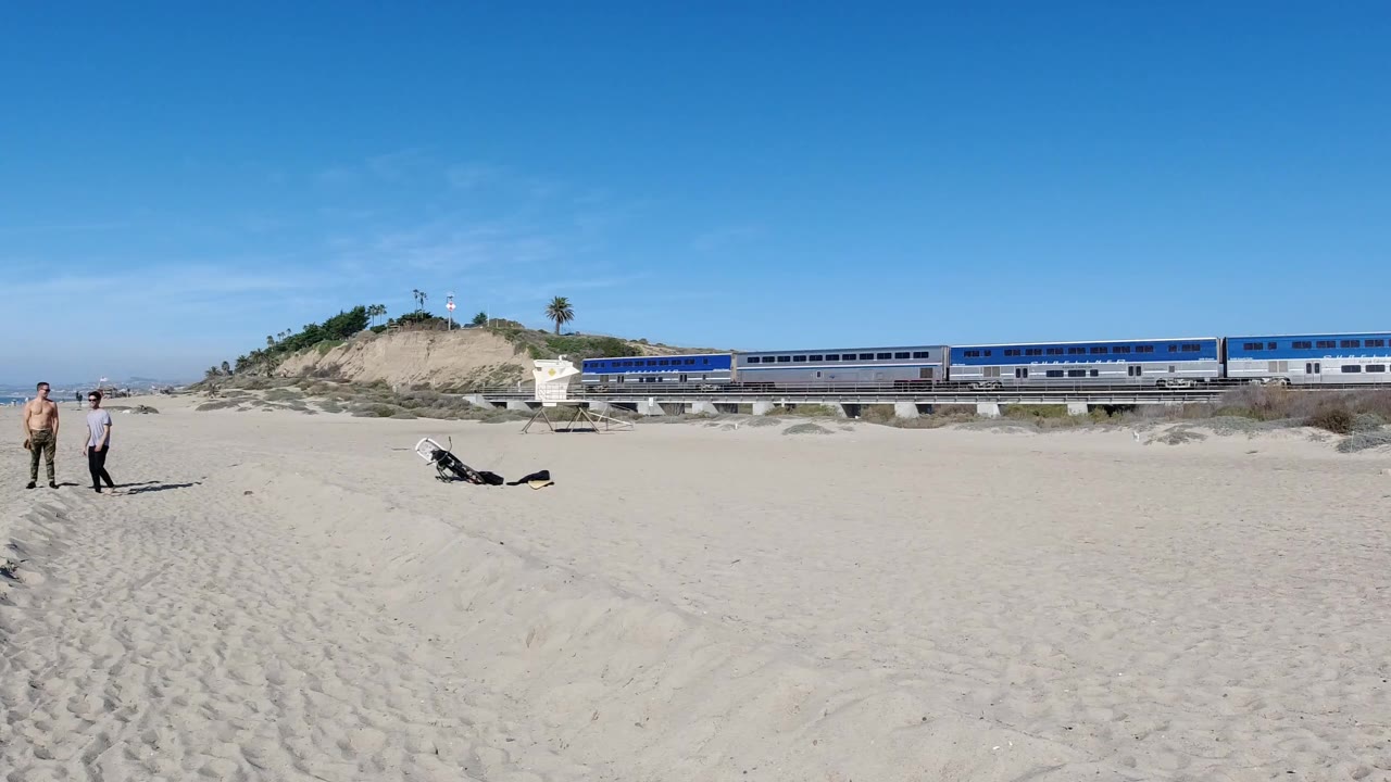 At Trestles Beach, California, a wide sandy foreground leads to two standing adults, a person lying on the sand, bicycles, a small lifeguard structure, and a blue and silver train behind the dunes.
