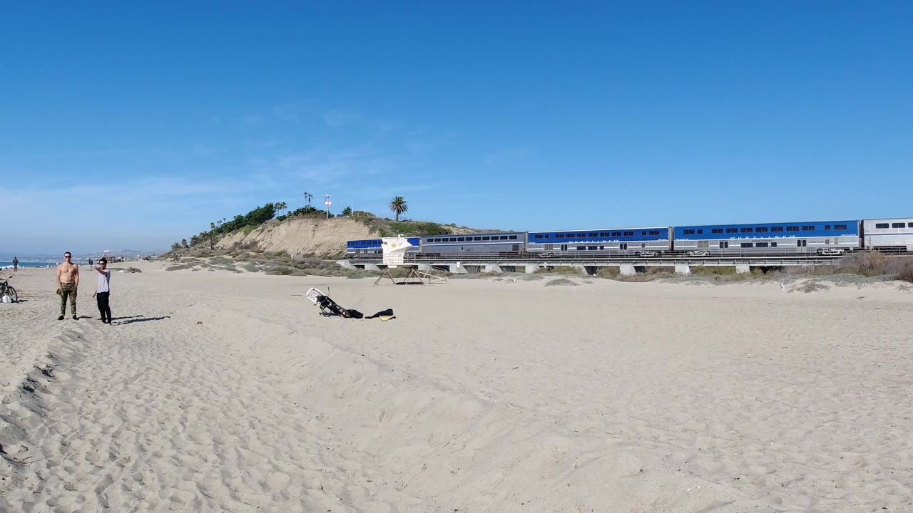 At 0:02, the blue and silver train continues across the dune-backed rail line, with two adults standing on the sand, bicycles nearby, and a clear sky over Trestles Beach, California.