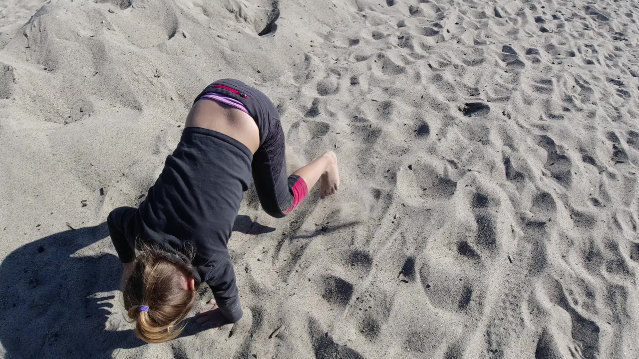At Trestles Beach, California, a young girl in dark clothing leans forward on rippled sand under bright sun.