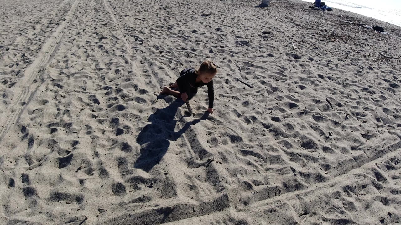 At Trestles Beach, California, a young girl kneels near the left side of a sandy frame with footprints, track marks, and a long shadow extending across the beach.