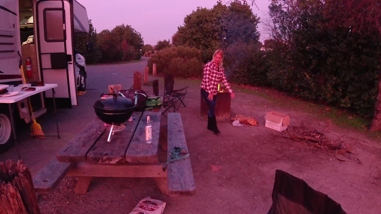 At Trestles Beach, California, the blond woman walks forward from the fire area, with the RV door edge, picnic table, grill, and roadside shrubs behind her.