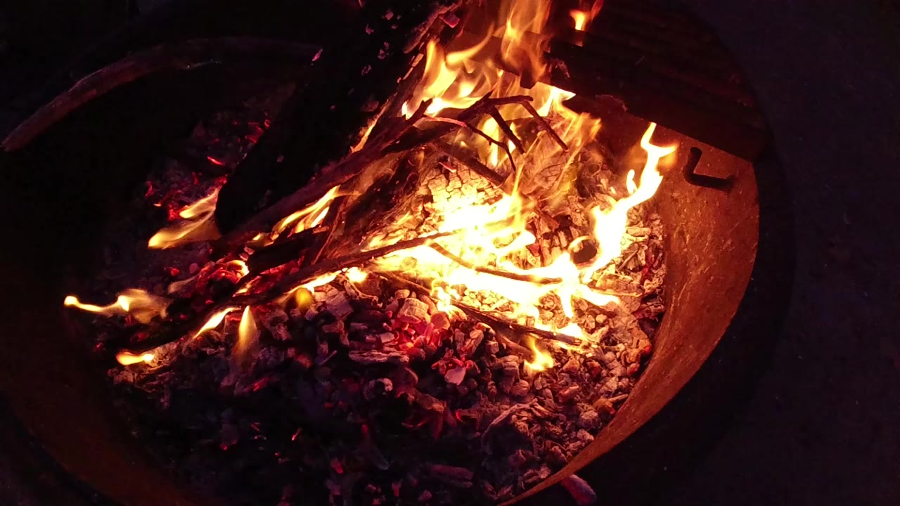 Round corrugated metal fire bucket at Trestles Beach, California, holding actively burning charred logs with bright yellow-orange flames in the upper portion and a red-orange ember bed at the base; no visible people.