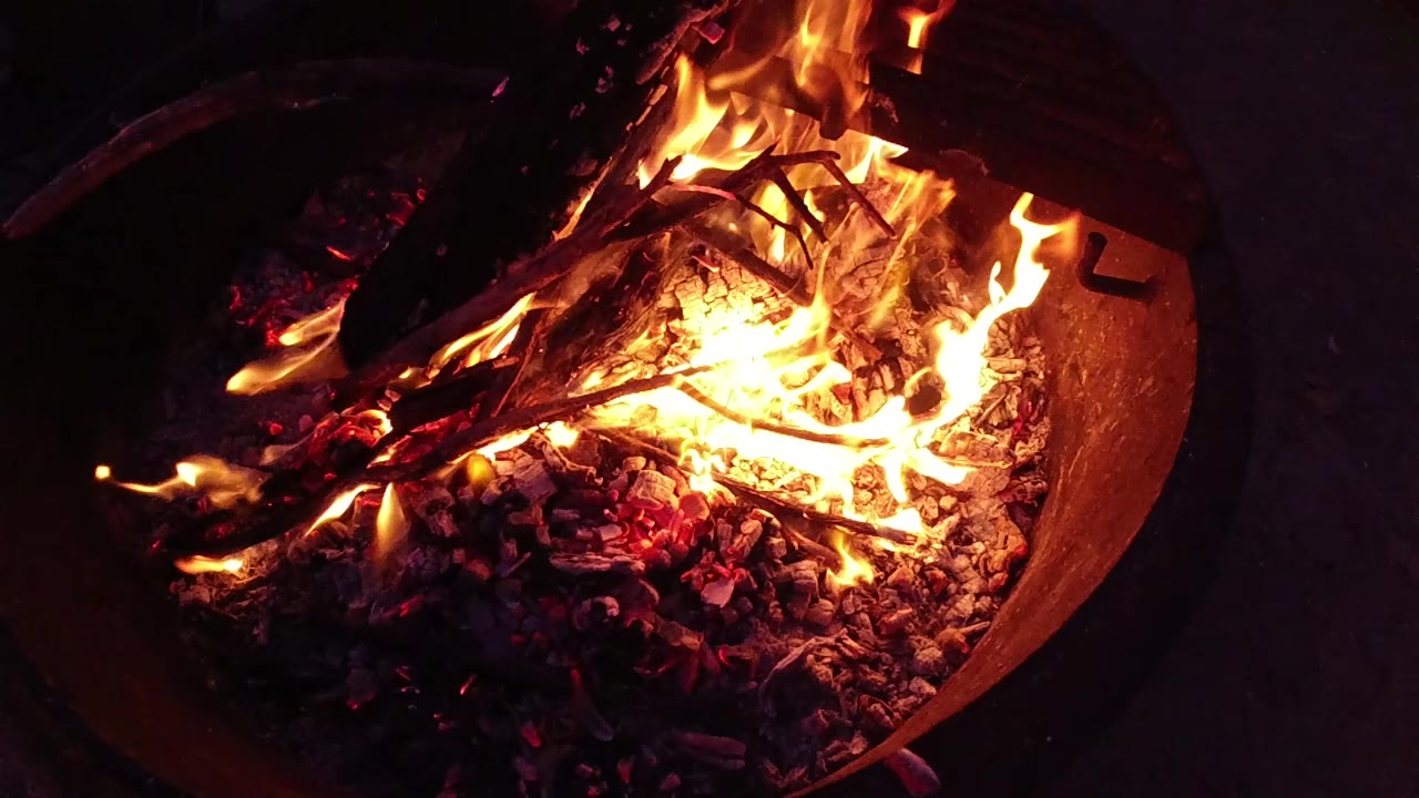 Corrugated metal fire bucket at Trestles Beach, California, showing active flames rising above charred branches and a bright ember bed, set against a black nighttime background; no visible people.