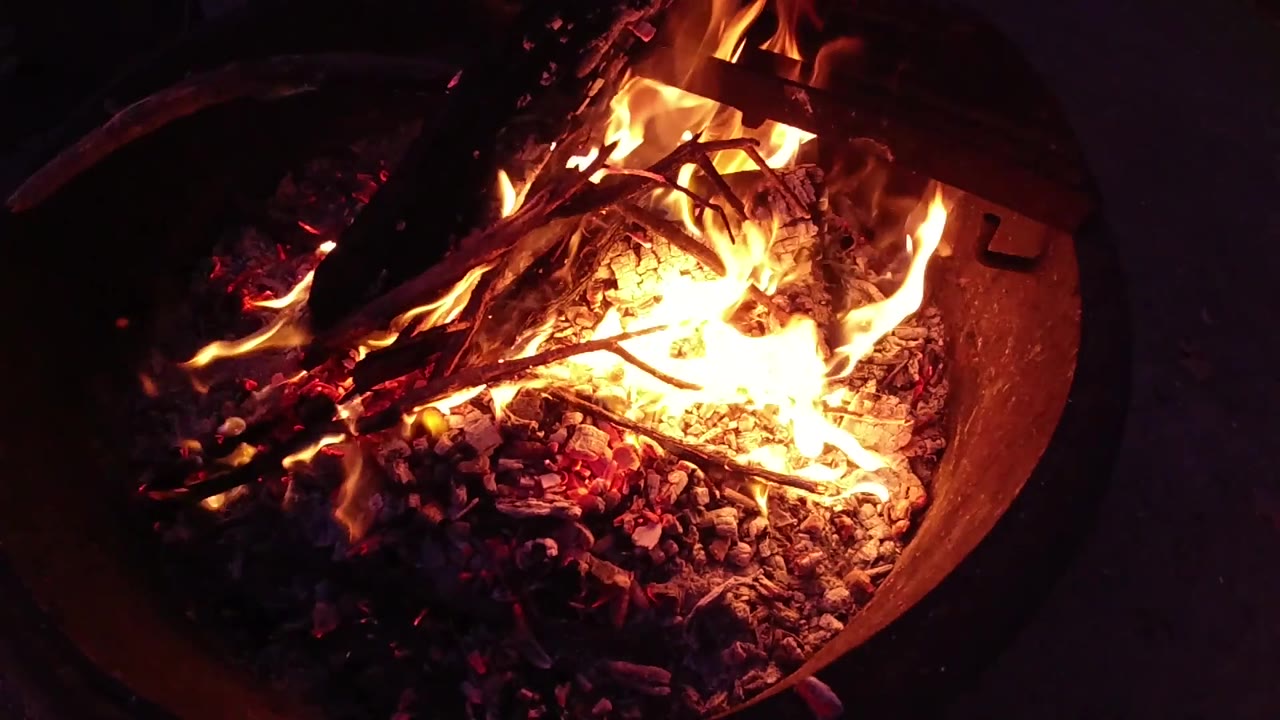 Close-up of round corrugated metal fire bucket at Trestles Beach, California, with active yellow-orange flames and a dense red-orange ember bed over charred wood; no visible people.