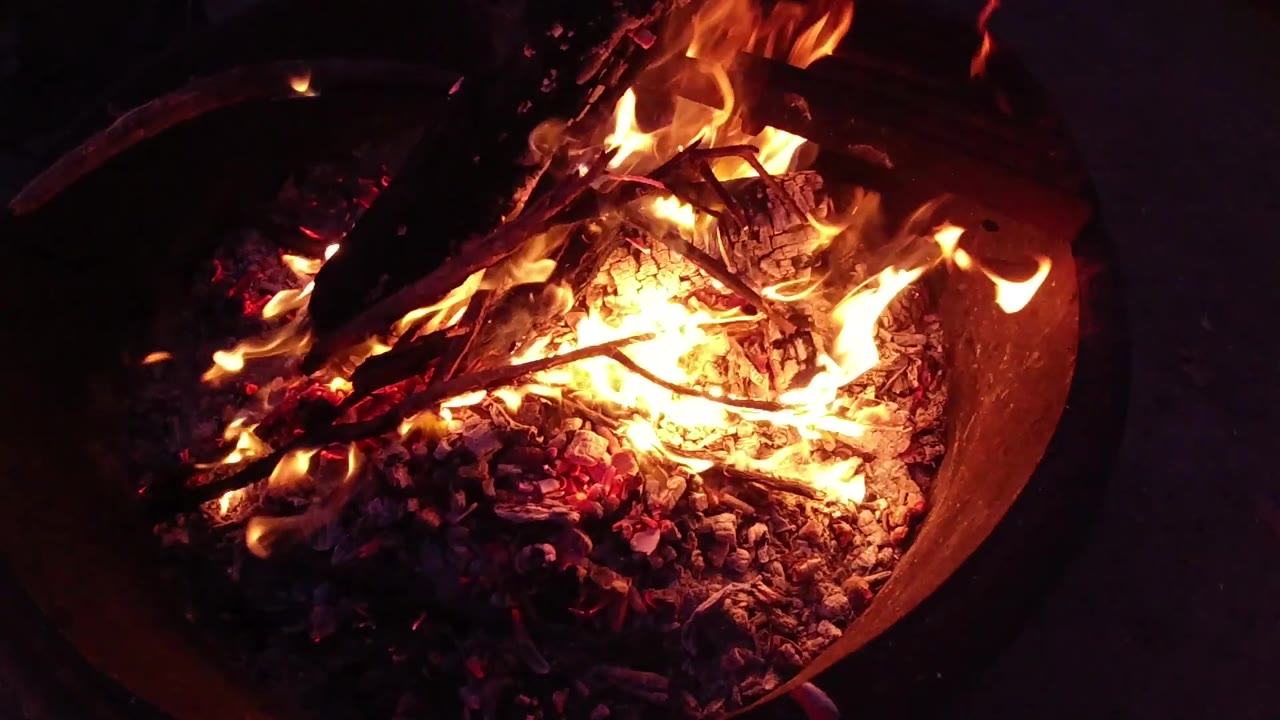 Round metal fire bucket at Trestles Beach, California, with bright yellow-orange flames concentrated toward the right side and charred branches spanning the full width of the container; no visible people.
