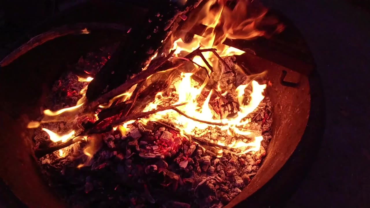 At Trestles Beach, California, bright flames rise over charred logs and glowing embers inside a round fire pit, with no visible people.